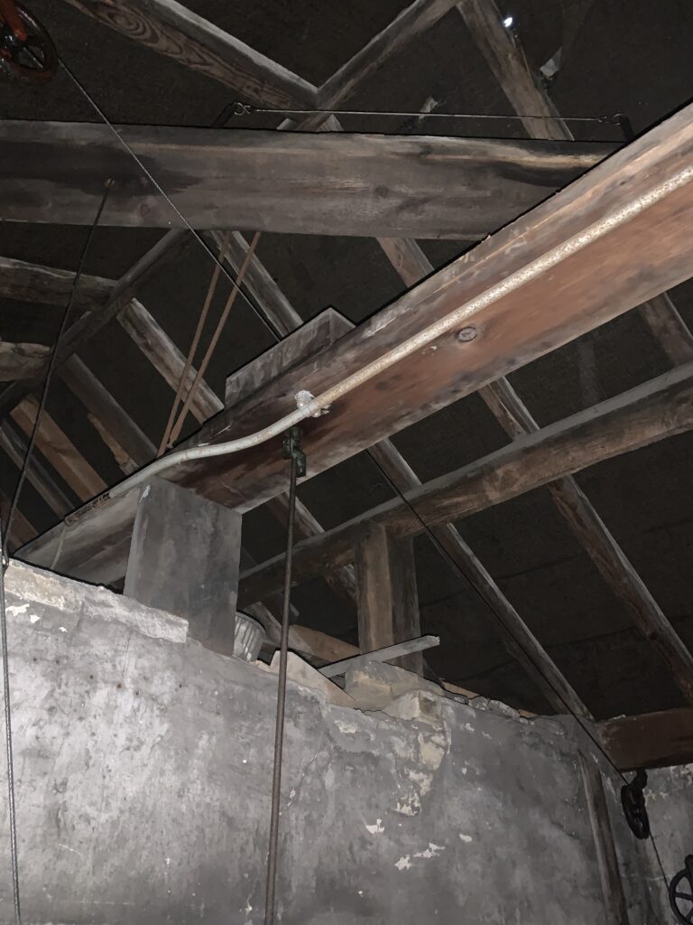 The ceiling of the clock tower. The bell wire leads between the clock and the bell. The beam is structural to the roof of the tower